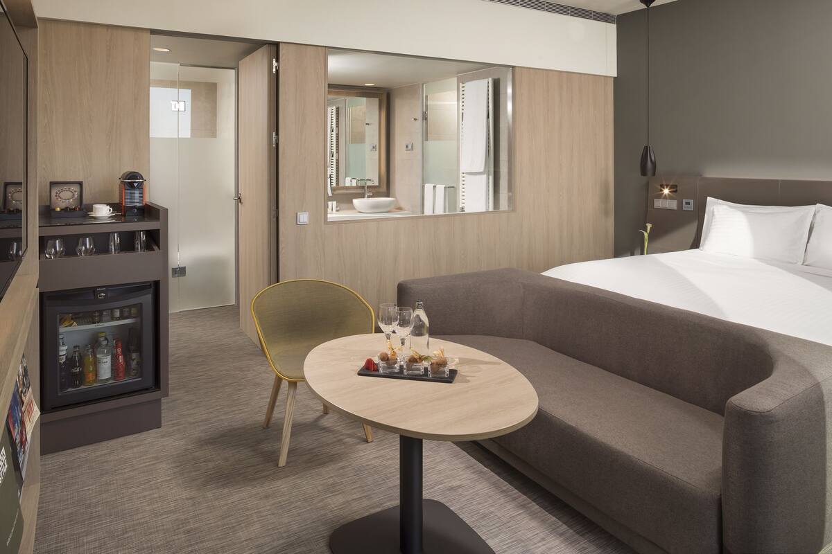 Quarto de hotel com cama, sofá, mesa pequena com copos e petiscos, minibar e espelho.