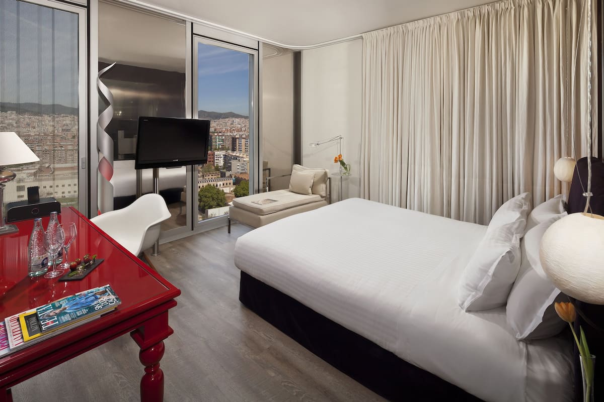 Quarto de hotel com uma cama grande, televisão, escrivaninha com mesa vermelha e vista para a cidade.