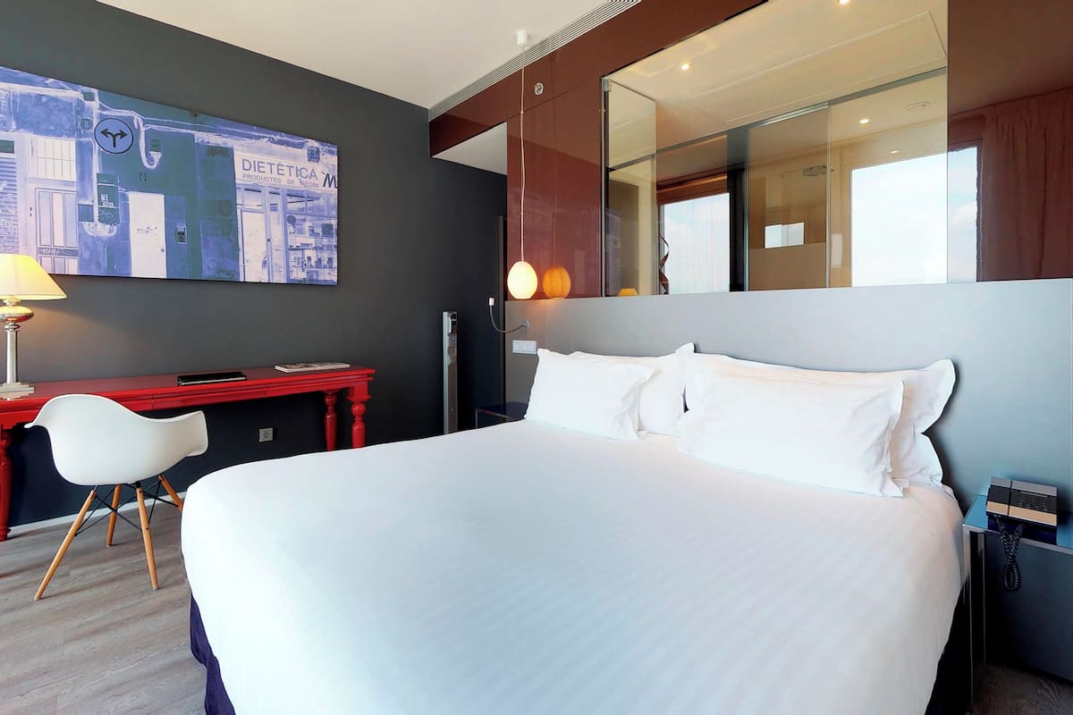 Quarto de hotel com uma cama grande, uma escrivaninha com cadeira, um abajur, um telefone e um espelho amplo.
