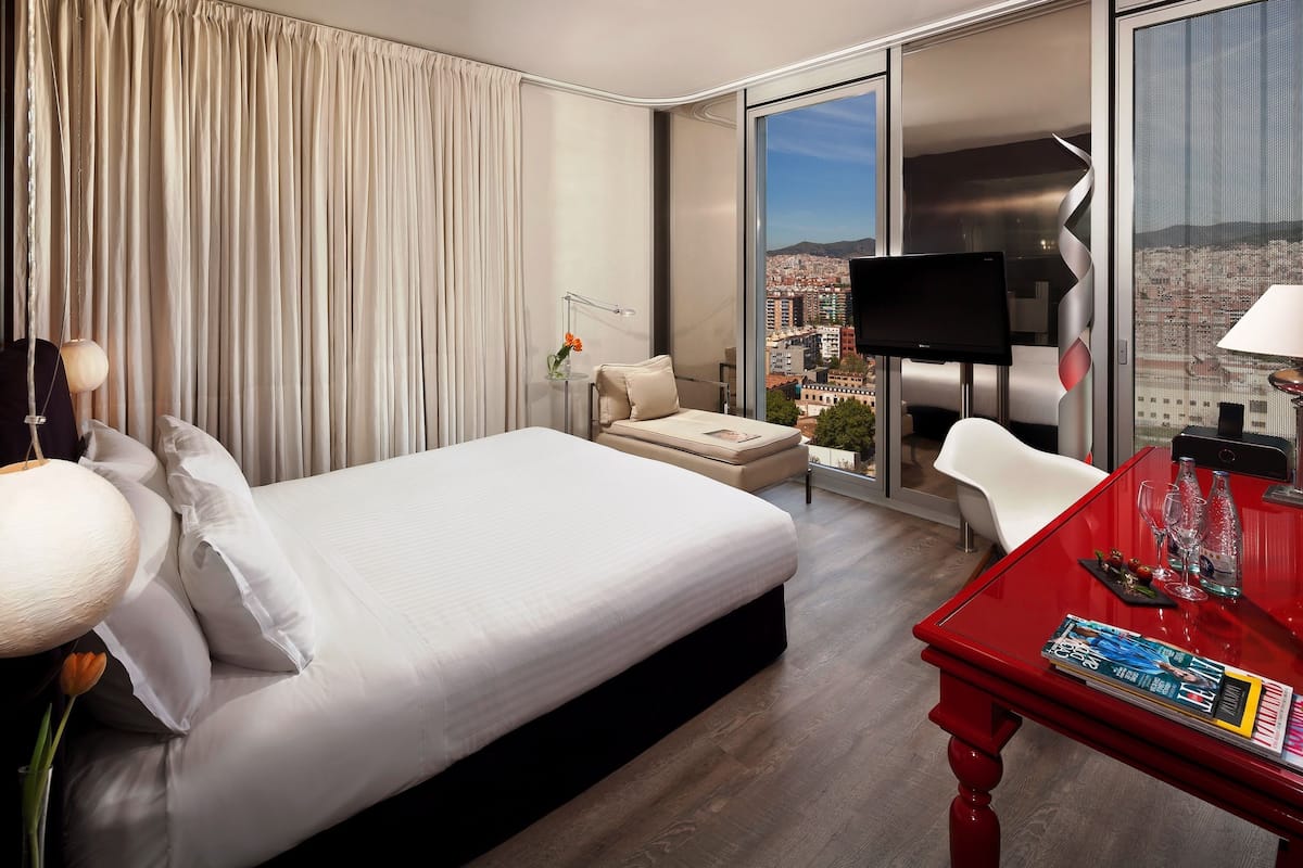 Quarto de hotel com cama, escrivaninha, poltrona, televisão e vista para a paisagem urbana através de grandes janelas.