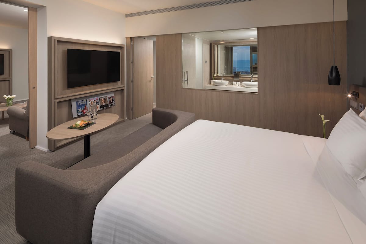 Quarto de hotel moderno com uma cama grande, televisão de tela plana, uma mesa pequena com revistas, um sofá e um banheiro com espelho grande e pia.