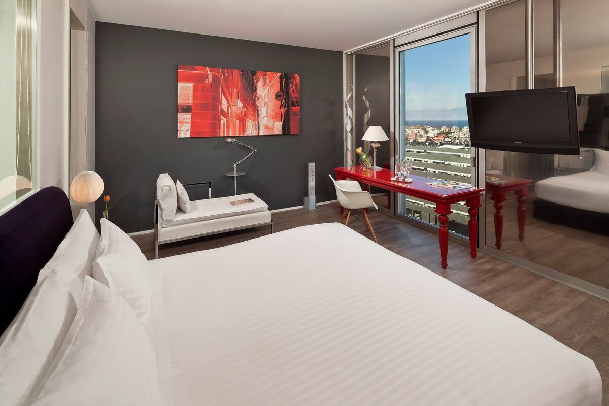 Quarto de hotel com cama, escrivaninha, cadeira, televisão, janela com vista para a cidade e um quadro na parede.