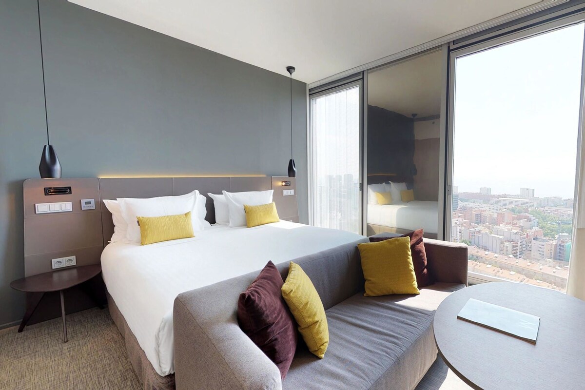 Habitación de hotel con una cama grande, un sofá, una mesita y vistas de la ciudad a través de un ventanal amplio.