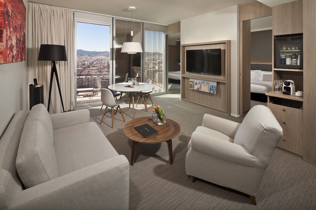 Quarto de hotel moderno com sofá, poltrona, mesa de jantar e televisão.