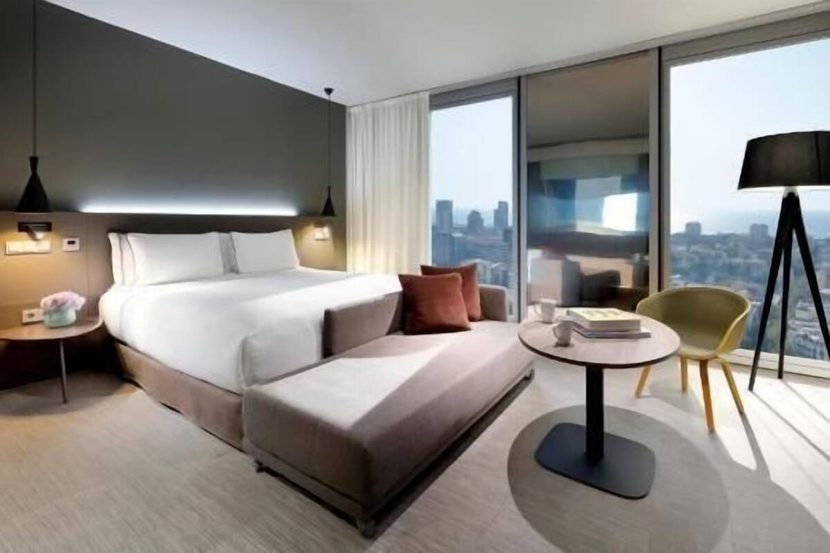 Quarto de hotel moderno com uma cama grande, um sofá, uma mesinha e vista para a cidade.