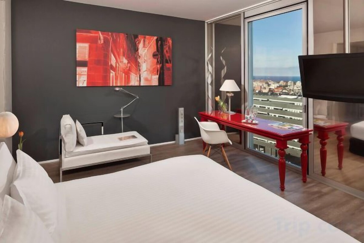 A hotel room with a bed, a desk, a chair, a TV, and a large window with a city view.