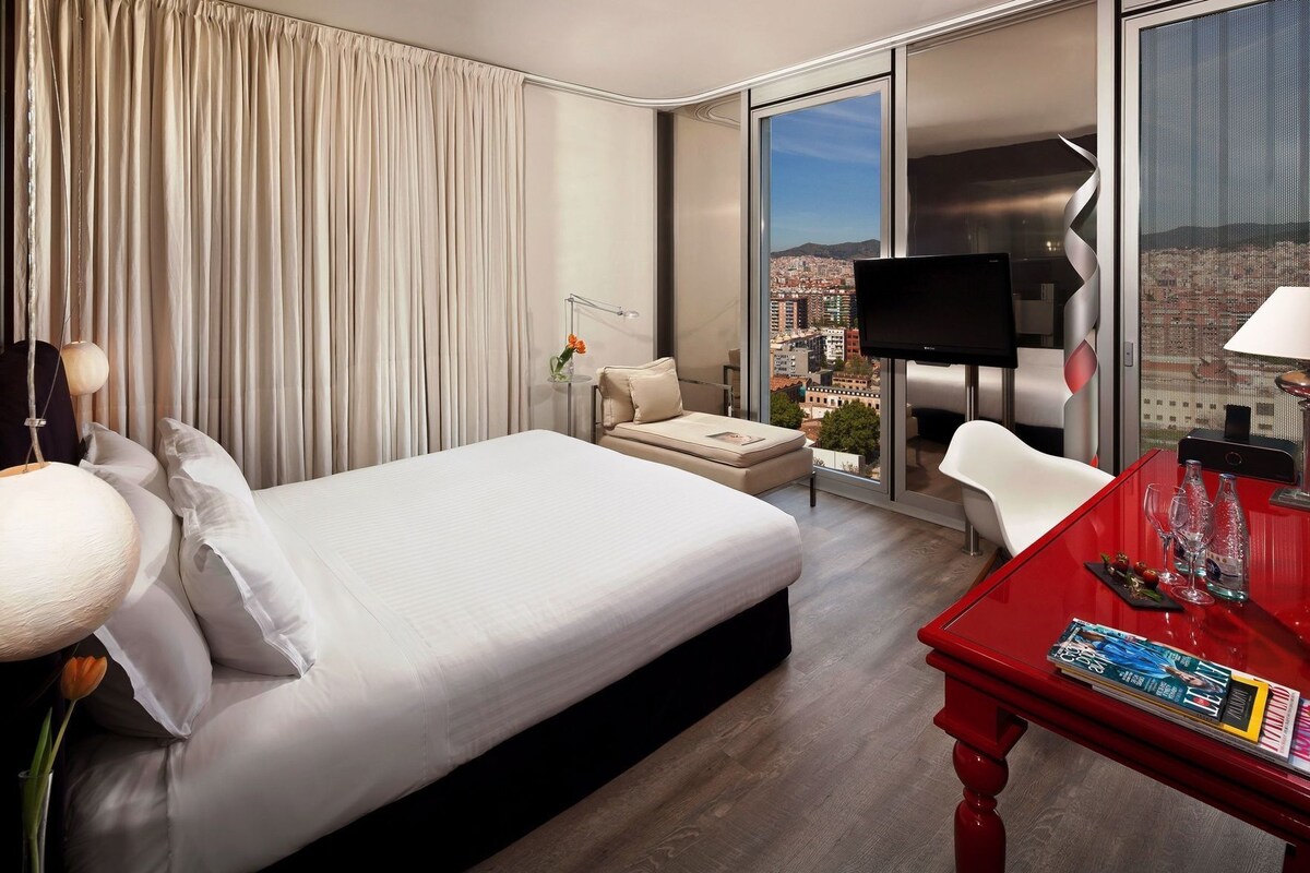 Habitación de hotel con cama, una mesa roja con revistas, televisión y vista a la ciudad.