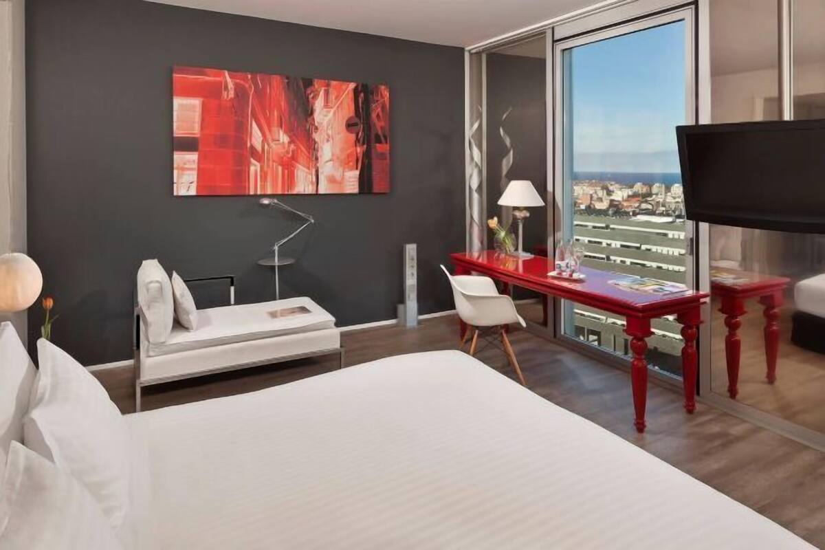 Quarto de hotel com cama, secretária, cadeira, sofá, televisão e uma grande janela com vista para a cidade.