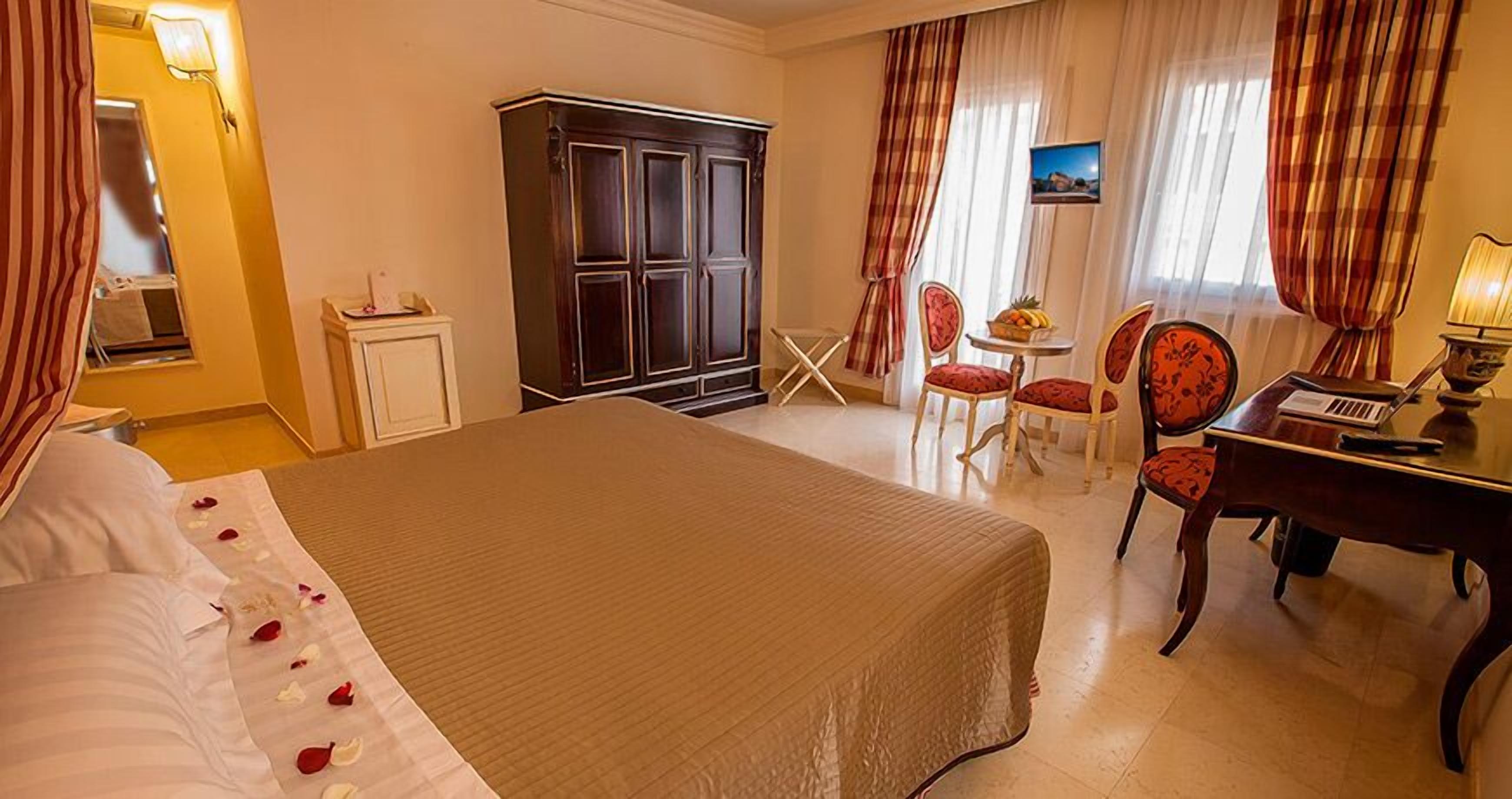 A hotel room with a large bed, a desk with a laptop, two chairs, a wardrobe, and a small table with fruit.