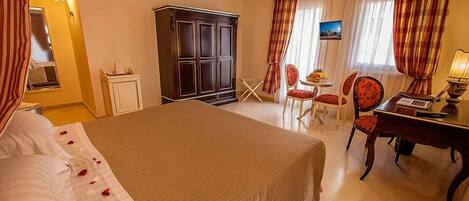 A hotel room with a large bed, a desk with a laptop, two chairs, a wardrobe, and a small table with fruit.