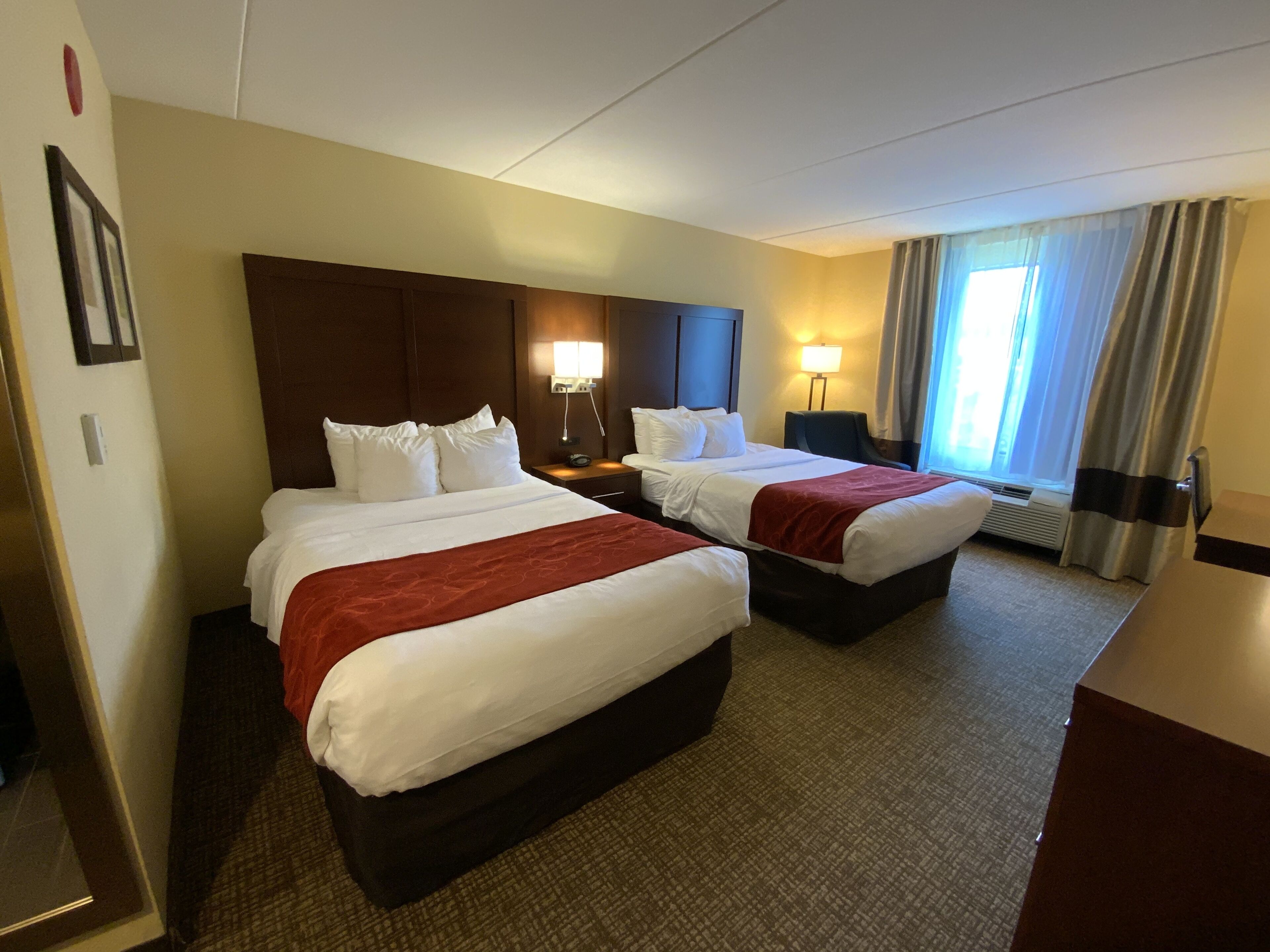 A hotel room with two beds, a nightstand, a lamp, a window with curtains, and a wall-mounted heater.