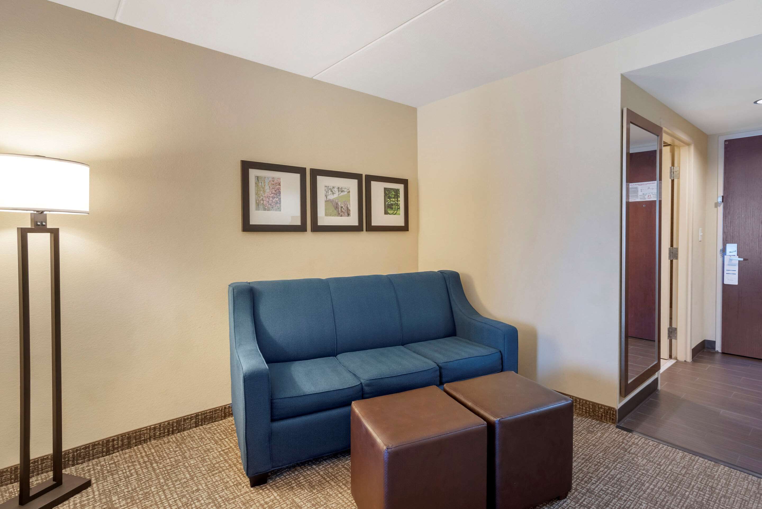 Habitació d'hotel amb un sofà blau, un otomà marró, una làmpada de peu i tres quadres a la paret.