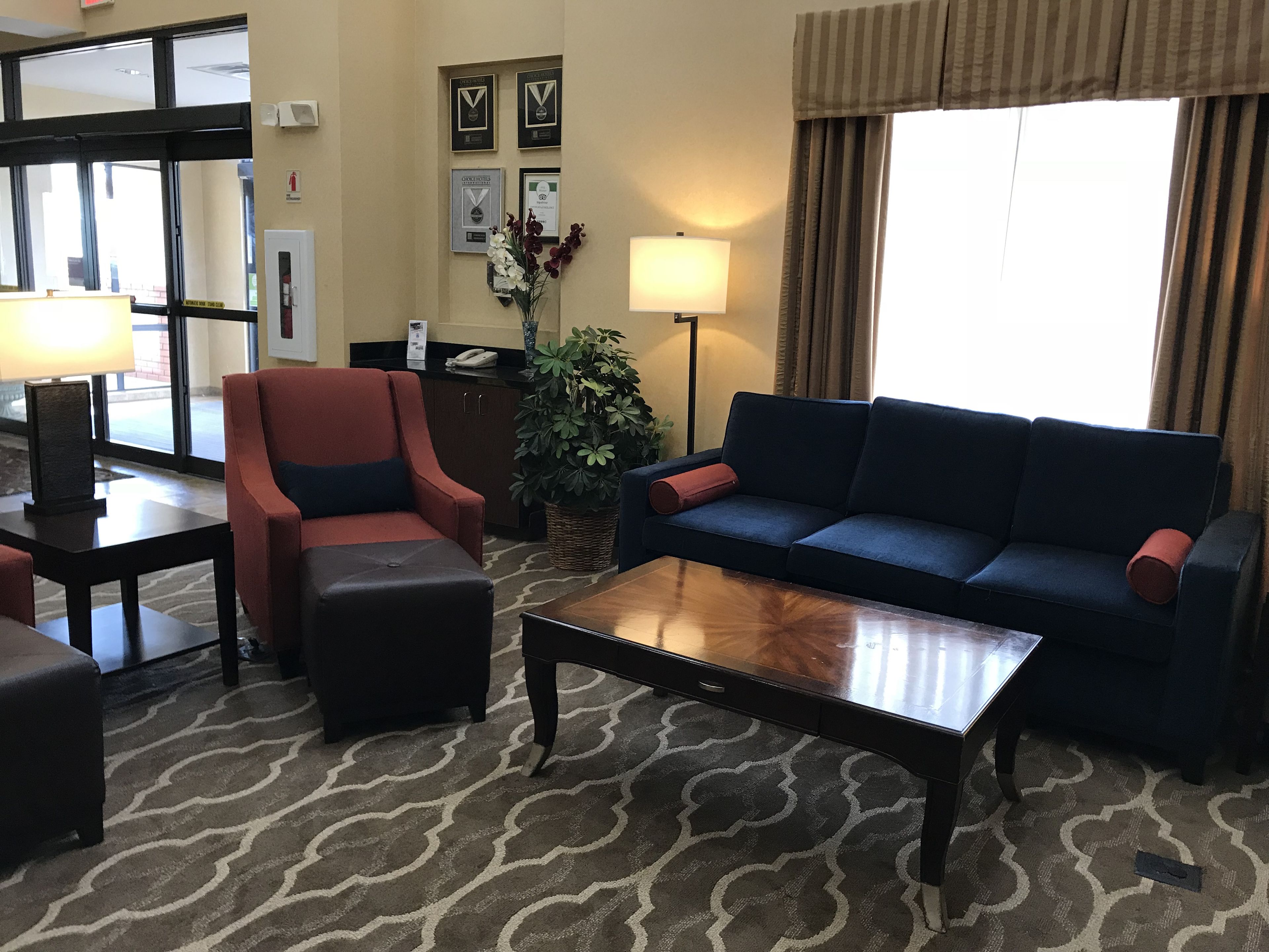 A hotel lobby with a blue sofa, red armchair, and a wooden coffee table. There is a reception desk with framed awards on the wall and a large window with curtains.