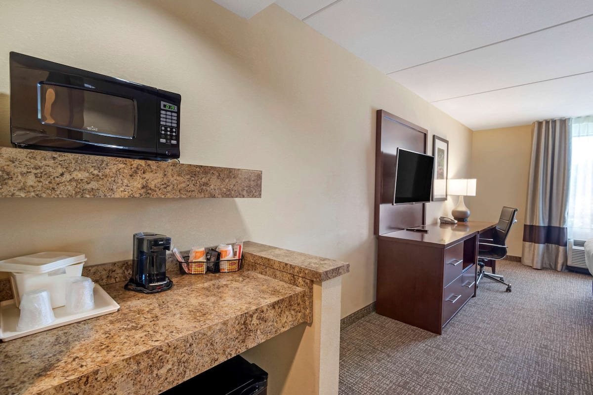 Habitación de hotel con kitchenette, que incluye microondas, cafetera y un refrigerador pequeño. Cuenta con un escritorio con silla, televisor y ventana con cortinas.
