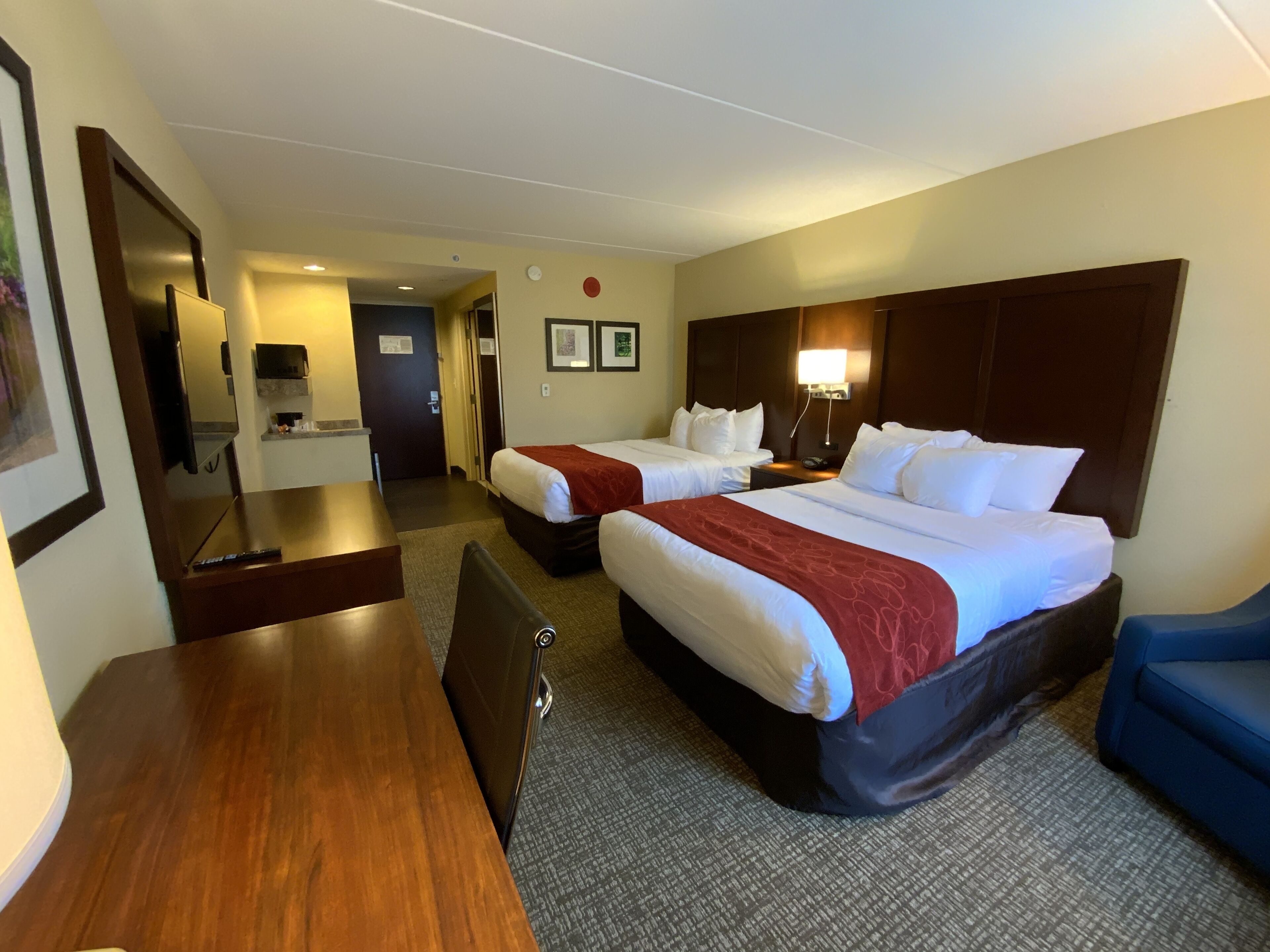 A hotel room with two beds, a desk, a chair, a TV, and a bathroom visible in the background.