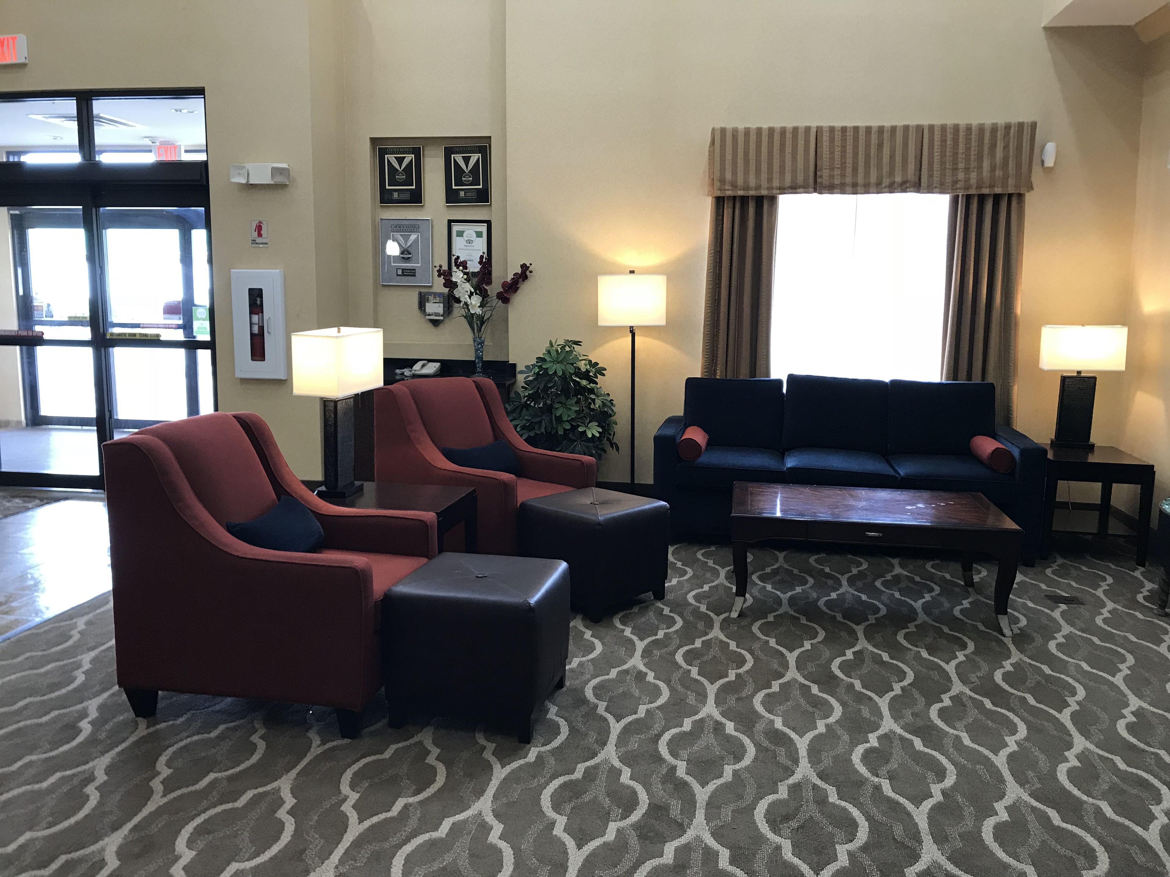 Un hall con mostrador de recepción, dos sillones, un sofá y una mesa de centro. En la pared hay cuadros con reconocimientos, una planta y una ventana con cortinas.