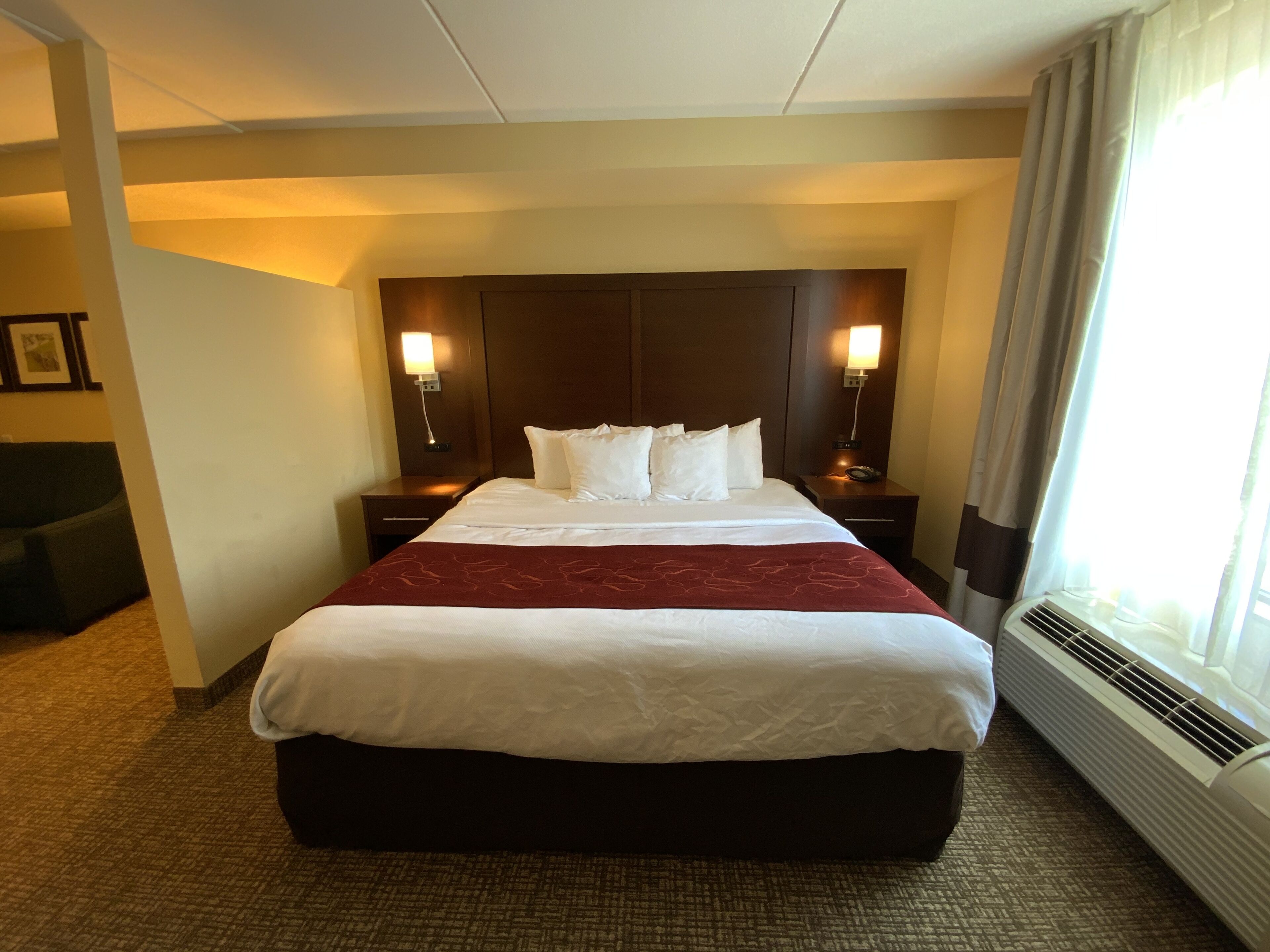 Una habitación de hotel con una cama grande, dos mesitas de noche con lámparas, un televisor montado en la pared y un ventanal con cortinas.