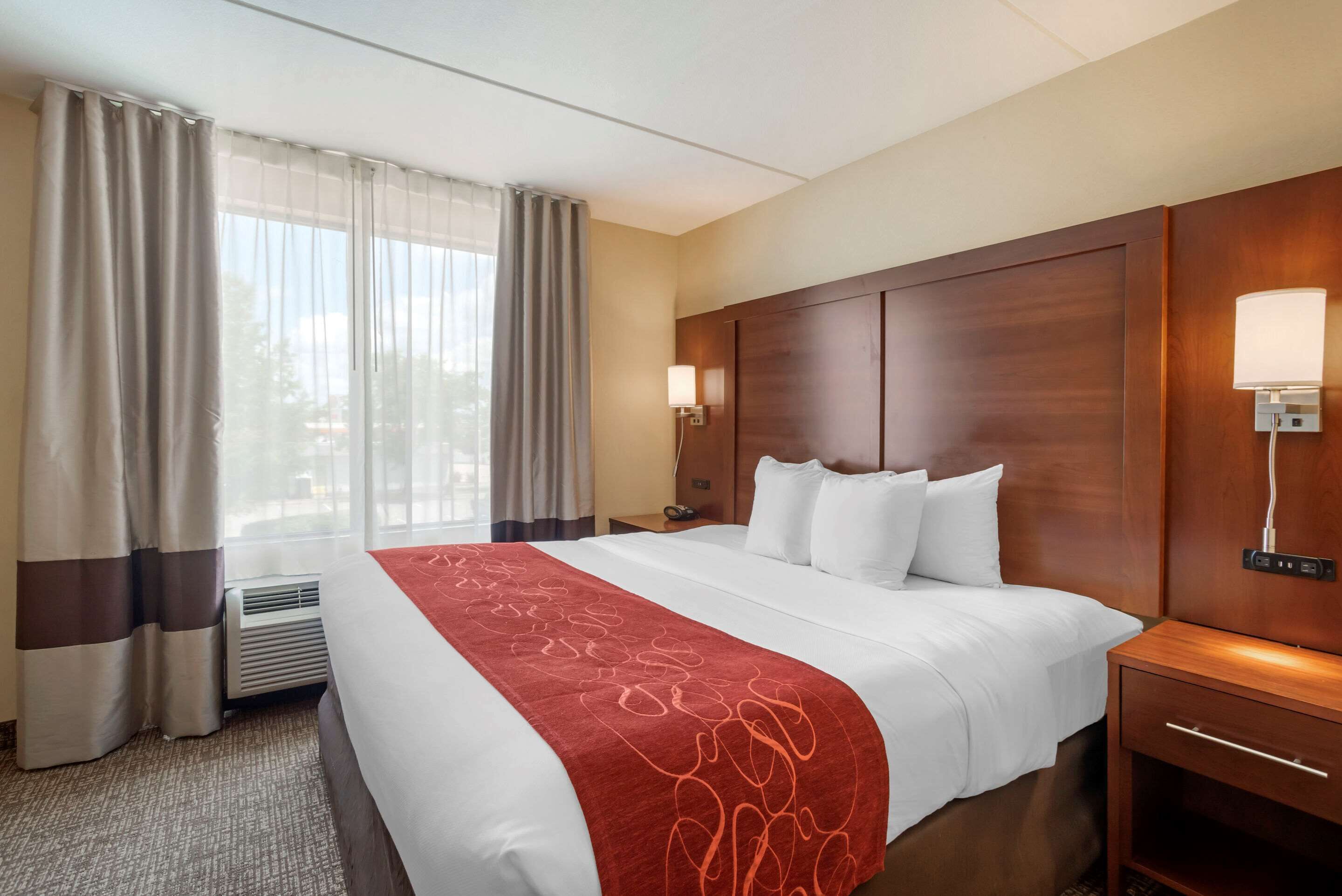 A hotel room with a large bed, a wooden headboard, a nightstand with a lamp, and a window with curtains.