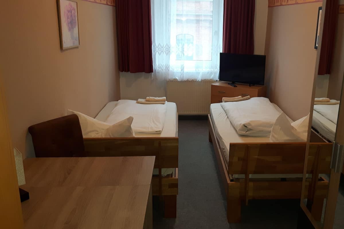 Ein Hotelzimmer mit zwei Einzelbetten, einem Holztisch, einem Fernseher auf einem Holzständer und einem Fenster mit Vorhängen.