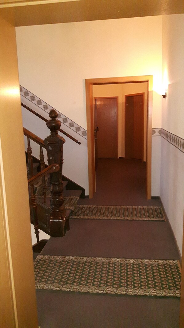 Un pasillo con piso alfombrado, una escalera de madera con barandilla y una puerta al final.