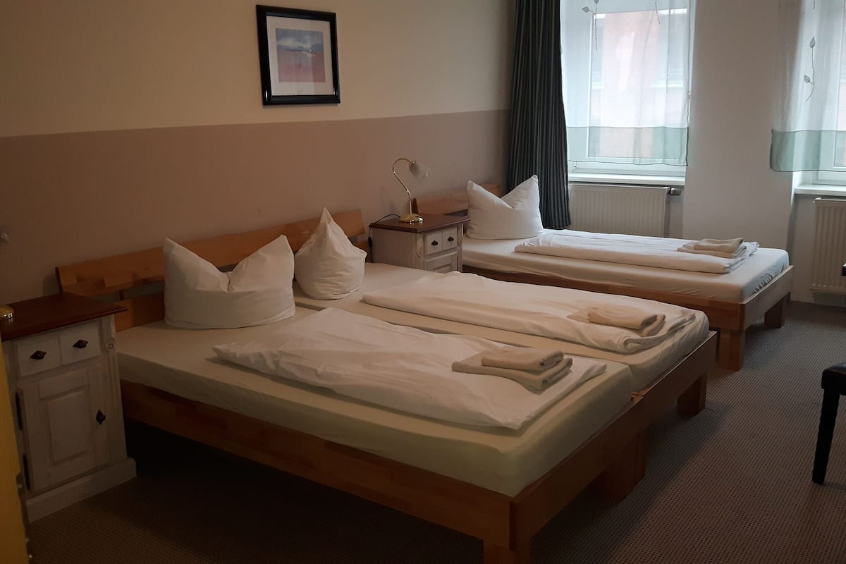 Ein Hotelzimmer mit zwei Einzelbetten, einem Nachttisch mit Lampe, einer kleinen Kommode und einem Fenster mit Vorhängen.