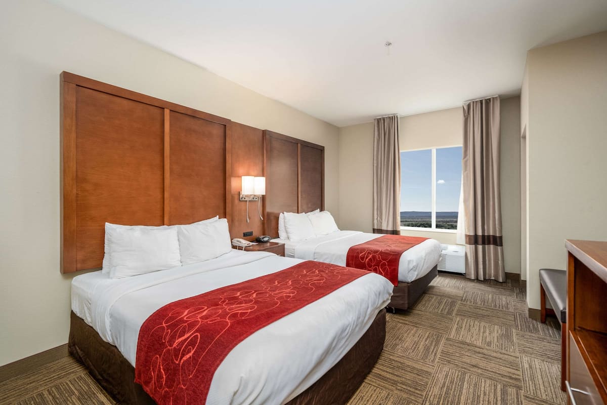A hotel room with two beds, a large wooden headboard, a nightstand with a lamp, and a view of the outdoors through a window.