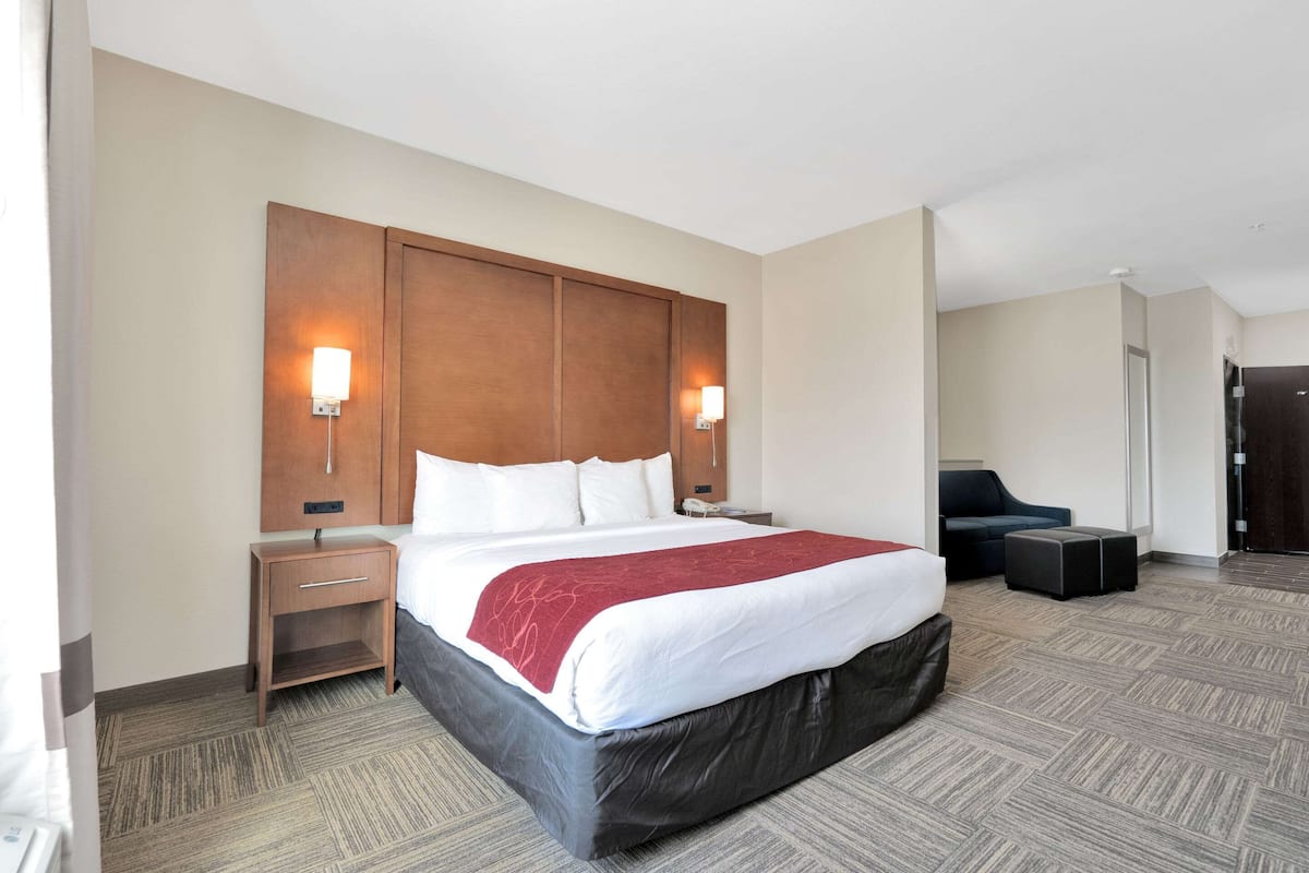 A hotel room with a large bed, a wooden headboard, two bedside tables, a nightstand, a desk, a chair, and a small ottoman.