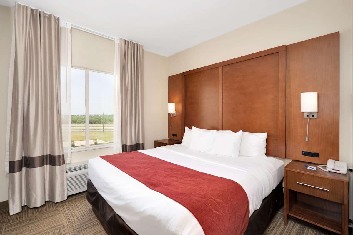 A hotel room with a large bed, two bedside tables, a nightstand, a window with curtains, and a view of the outdoors.