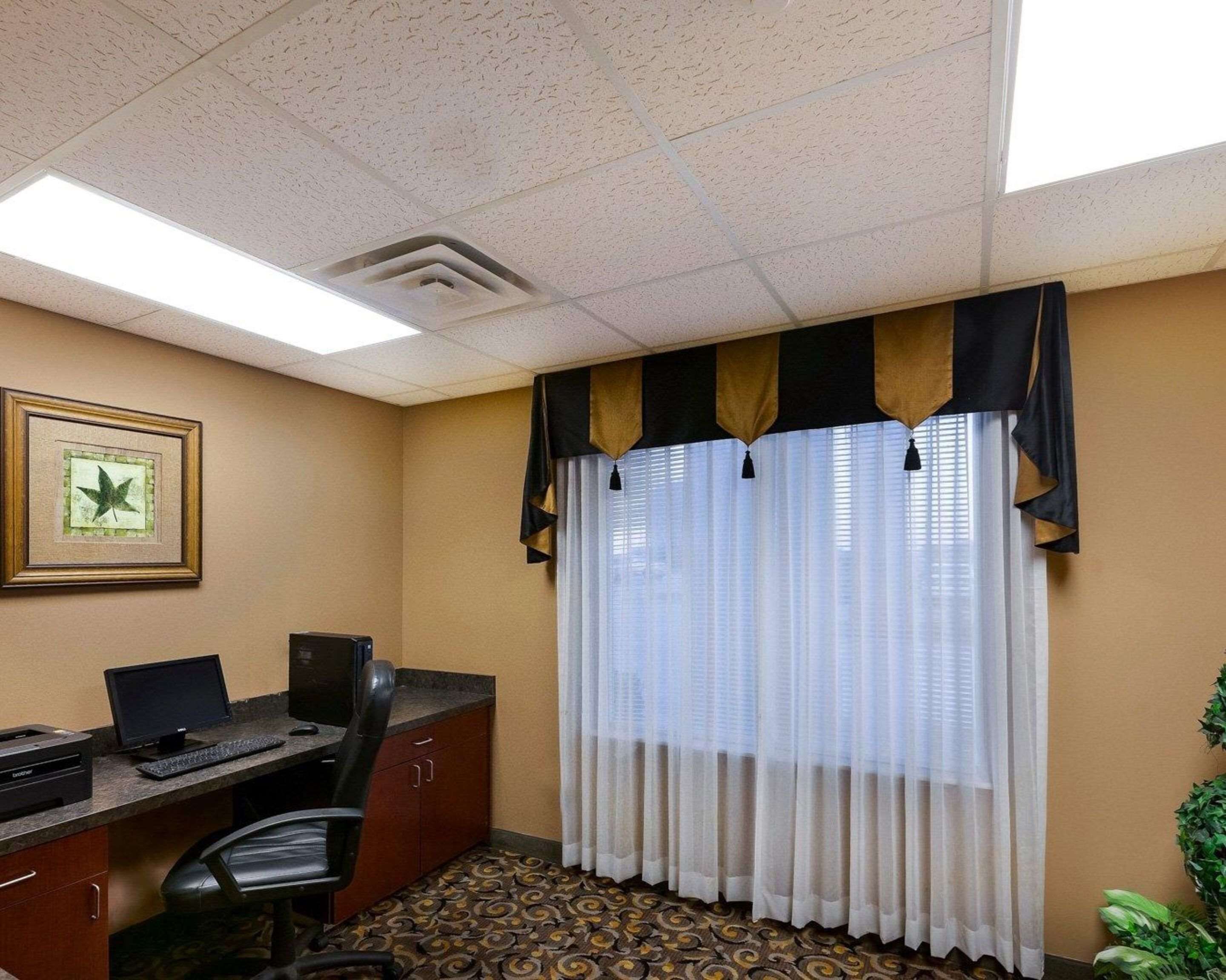 A hotel room with a desk, computer, and chair. There is a window with curtains and a plant on the right side.
