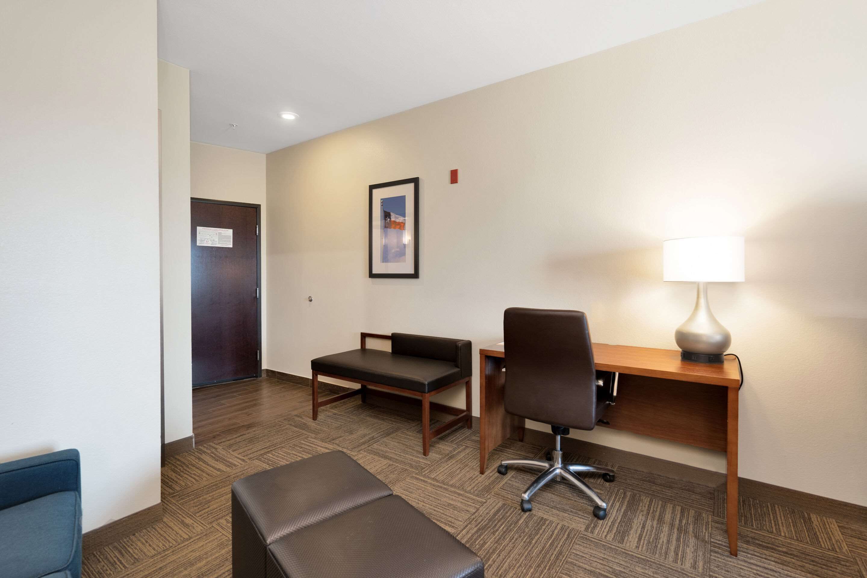A hotel room with a desk, chair, two ottomans, a lamp, and a framed picture on the wall.