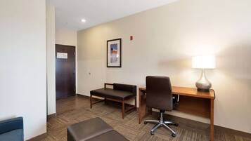 A hotel room with a desk, chair, two ottomans, a lamp, and a framed picture on the wall.