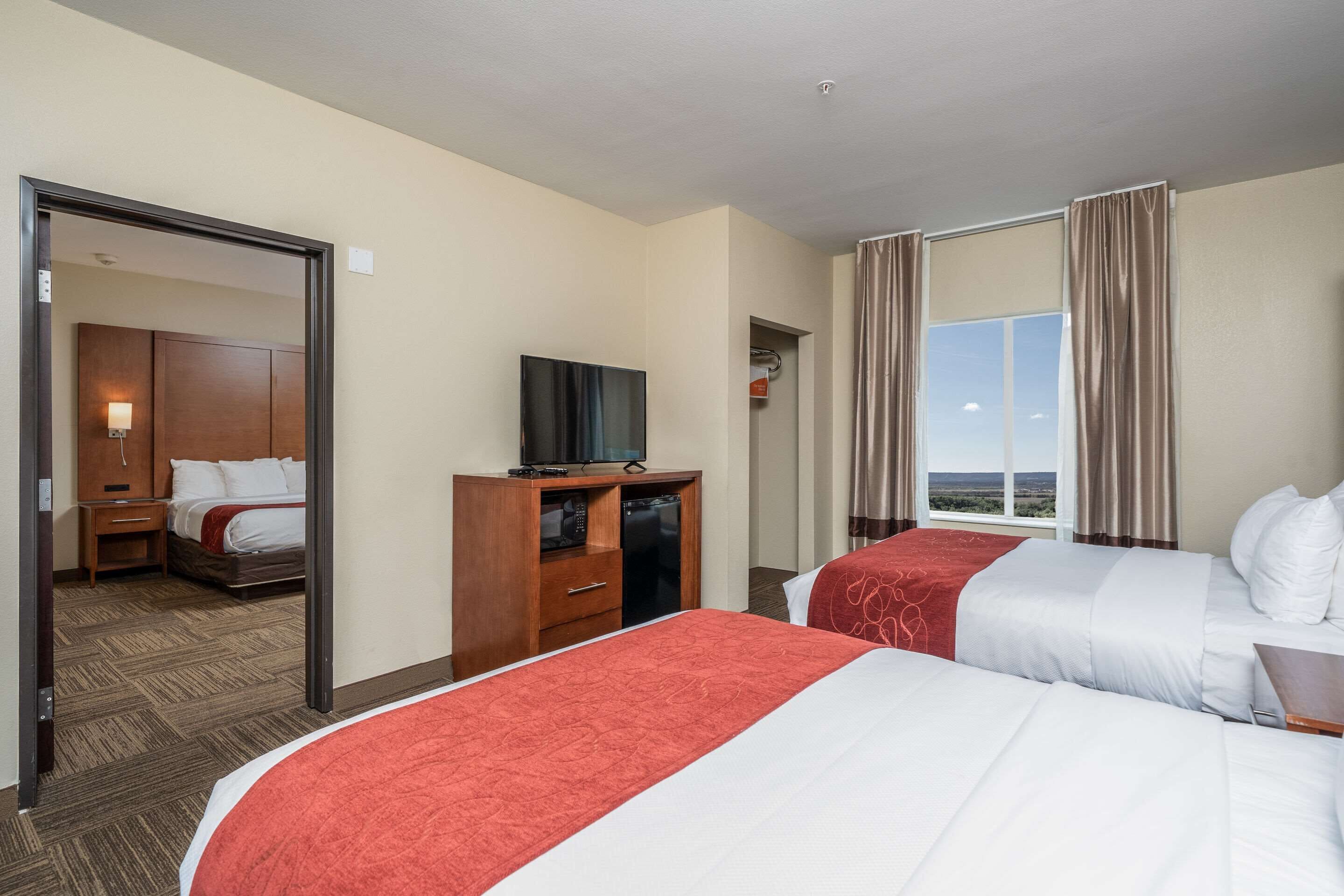 Quarto de hotel com duas camas, televisão sobre um móvel de madeira, espelho e janela com cortinas.