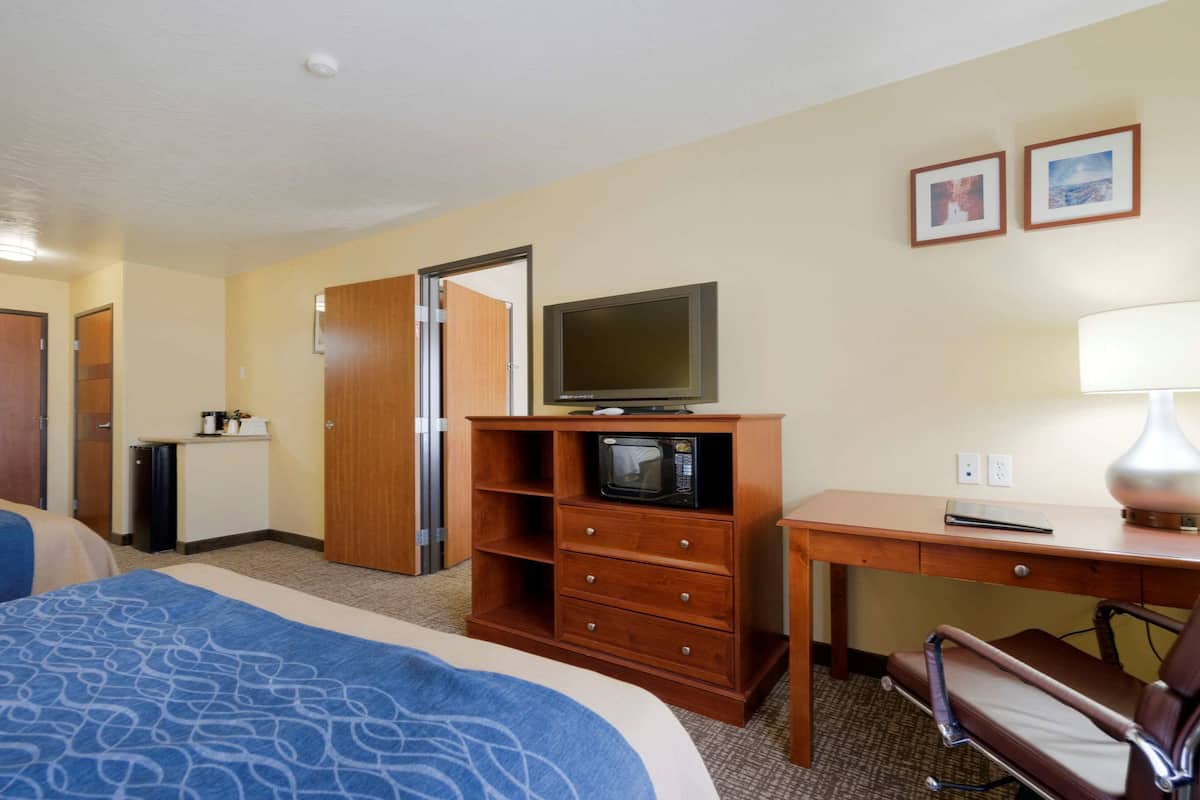 Habitación de hotel con cama, escritorio con lámpara, televisor sobre un mueble de madera y puerta que da a otra habitación.