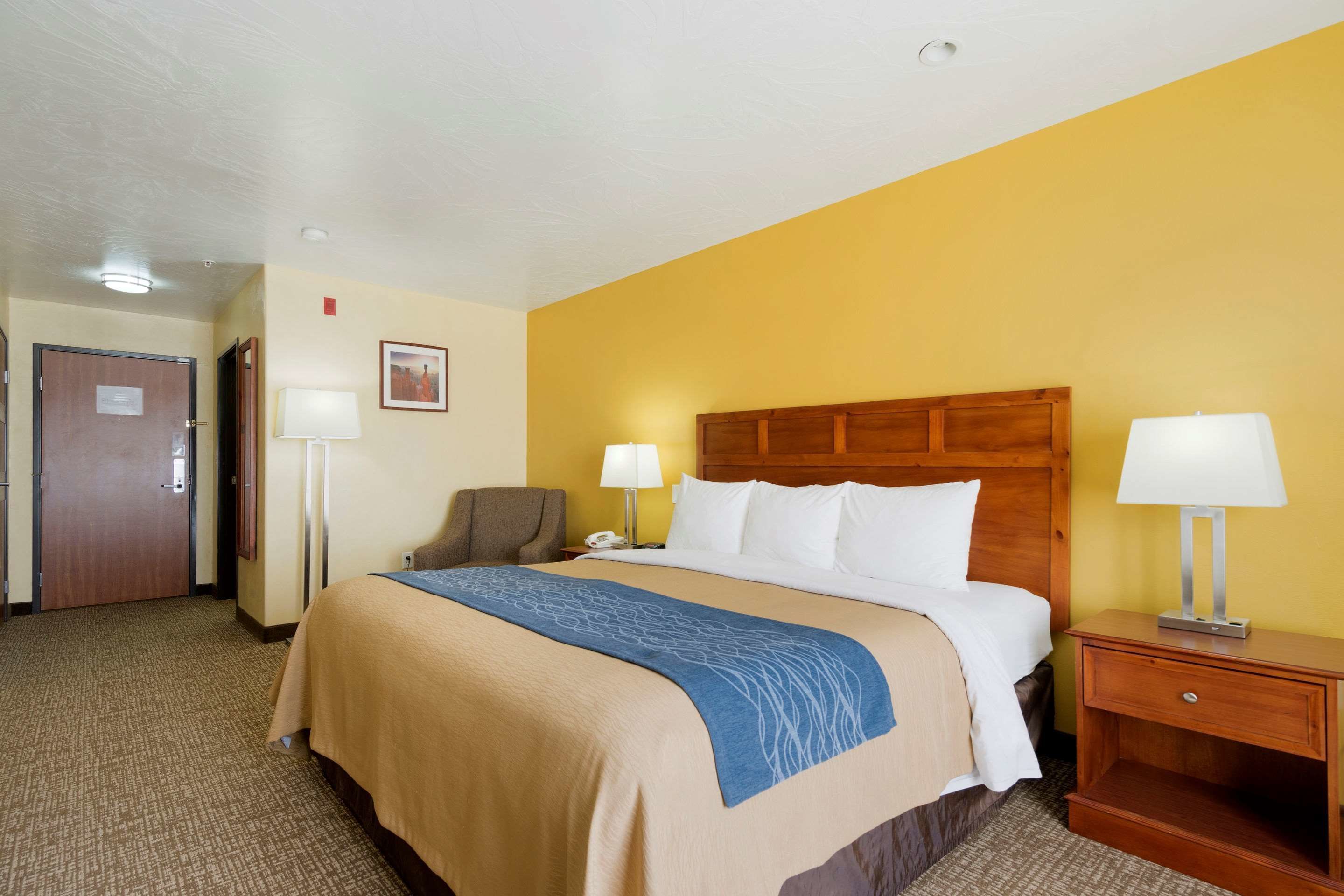 A hotel room with a bed, two bedside tables with lamps, a chair, a desk, and a door.
