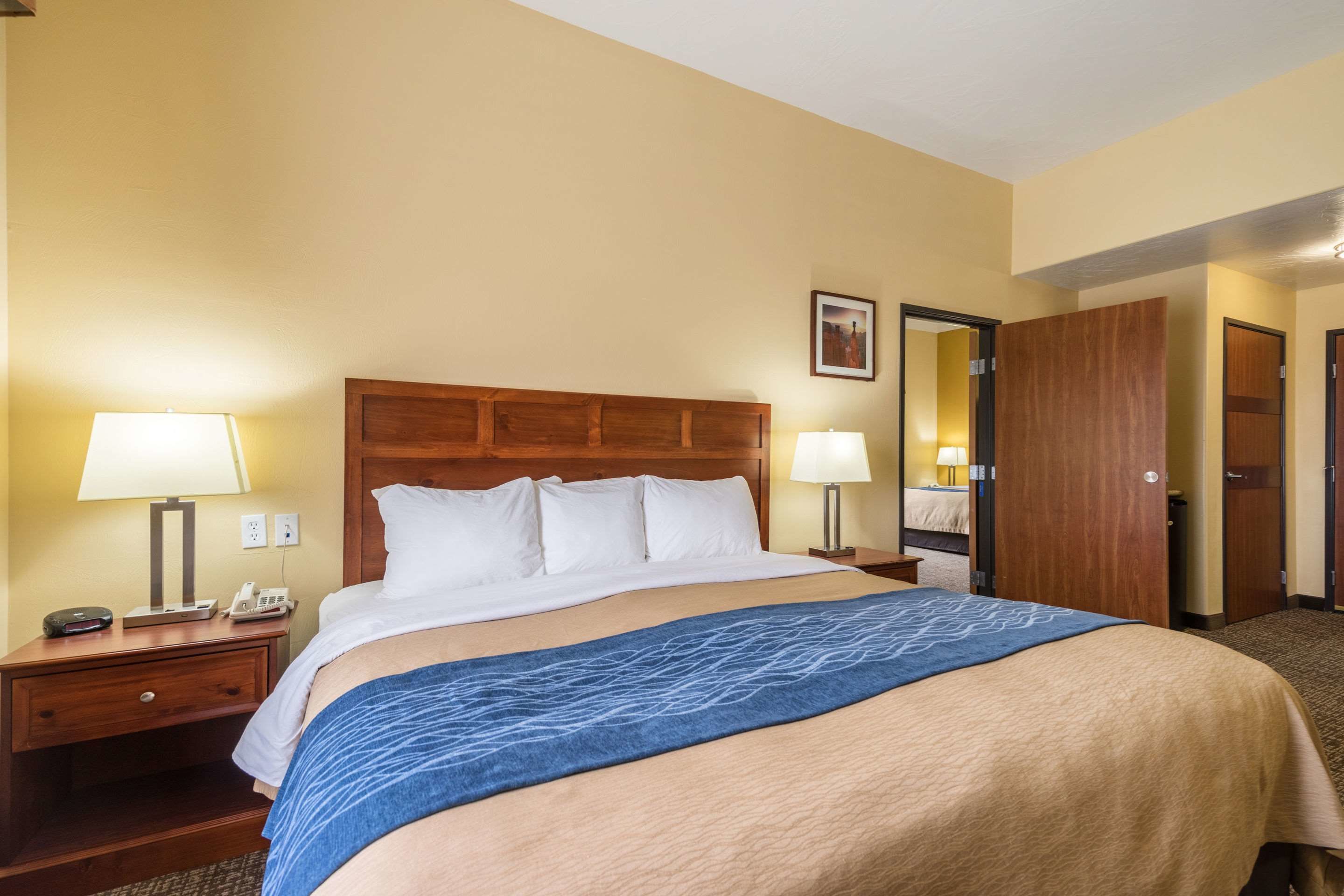 A hotel room with a large bed, wooden headboard, two bedside tables with lamps, a nightstand, and a door leading to another room.