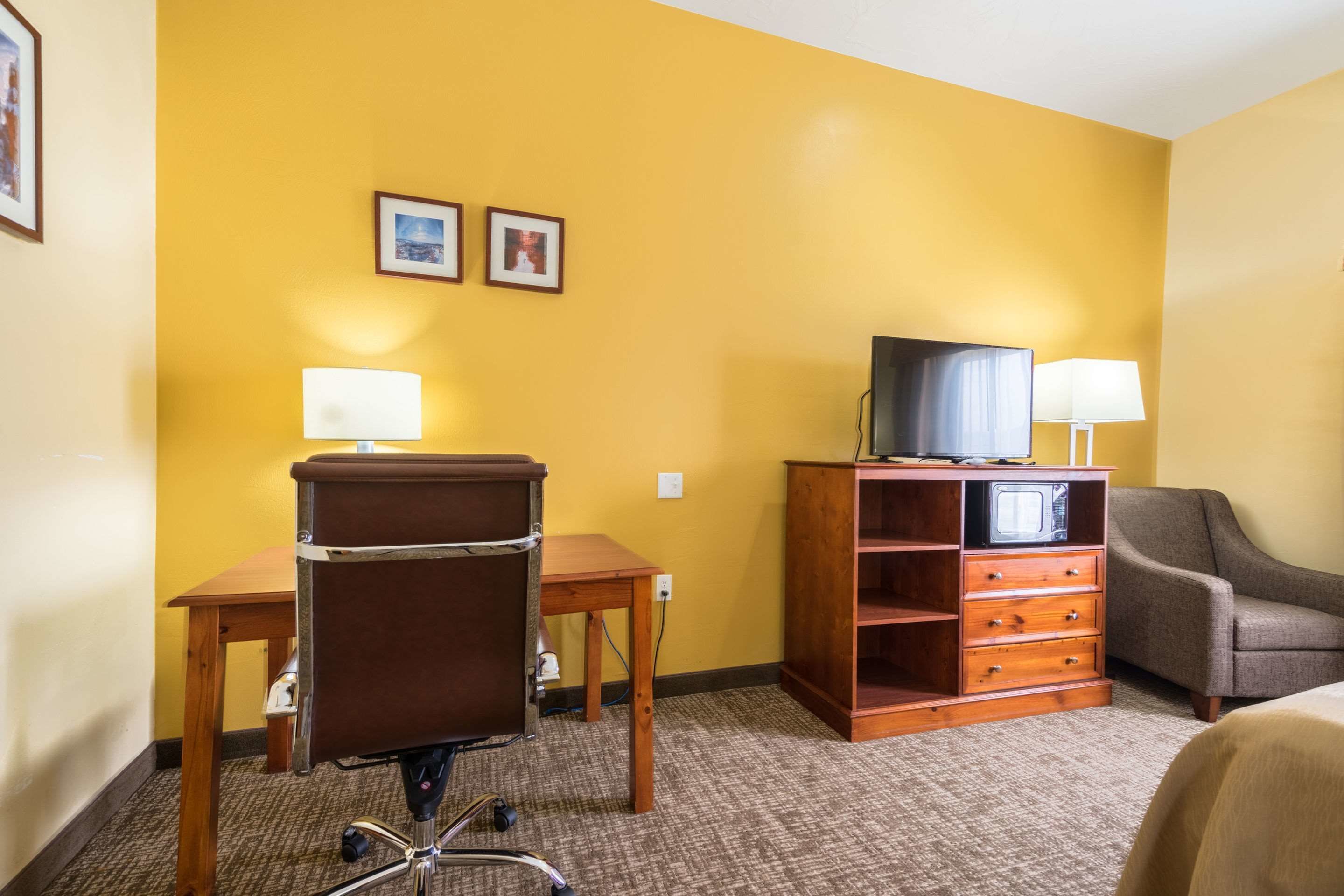 A hotel room with a brown desk, a brown office chair, a television on a wooden stand, a grey sofa, and two framed pictures on the wall.
