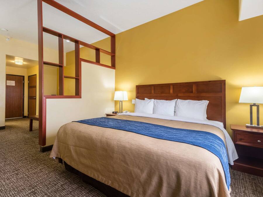 A hotel room with a large bed, wooden headboard, two bedside tables with lamps, a carpeted floor, and a door leading to another room.