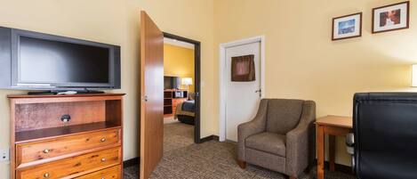 A hotel room with a flat-screen TV on a wooden dresser, a gray armchair, a small wooden table, and a desk with a chair.