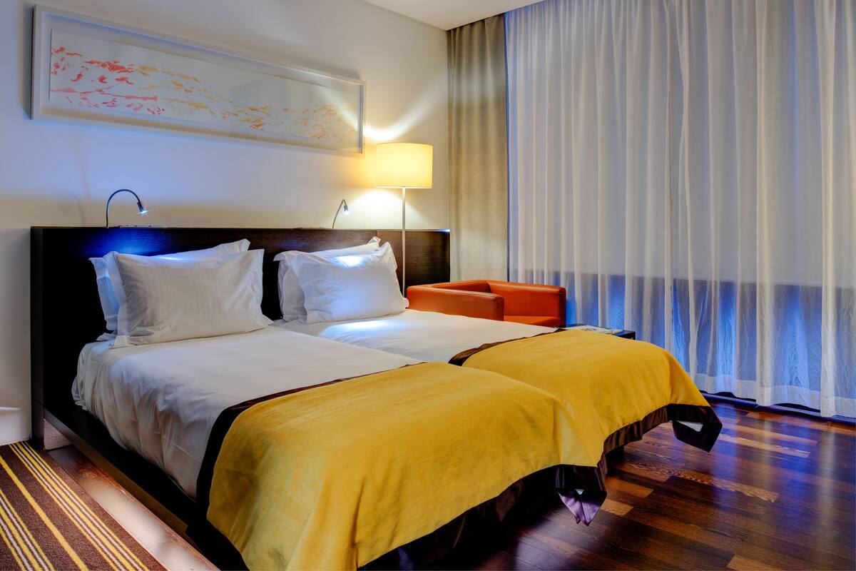 A double bed with white and yellow bedding, a wooden headboard, a bedside table with a lamp, and a view of a window with sheer curtains.