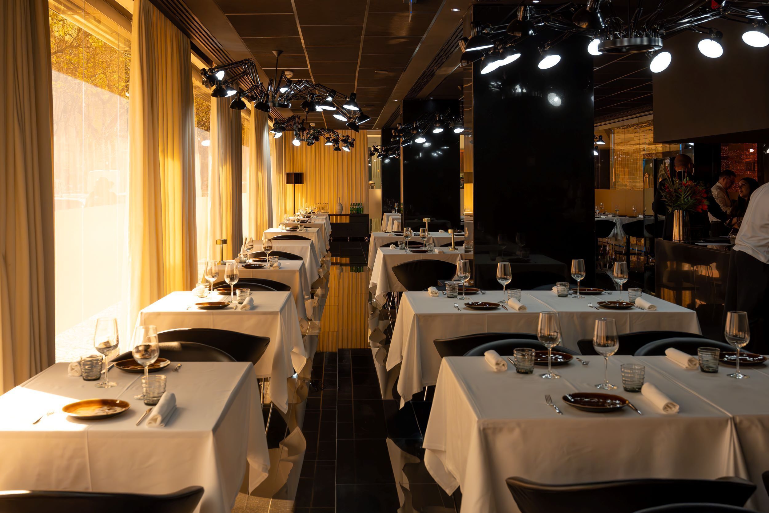 En restaurant med flere borde dækket til spisning, der har en lang disk og en mørk væg i baggrunden.
