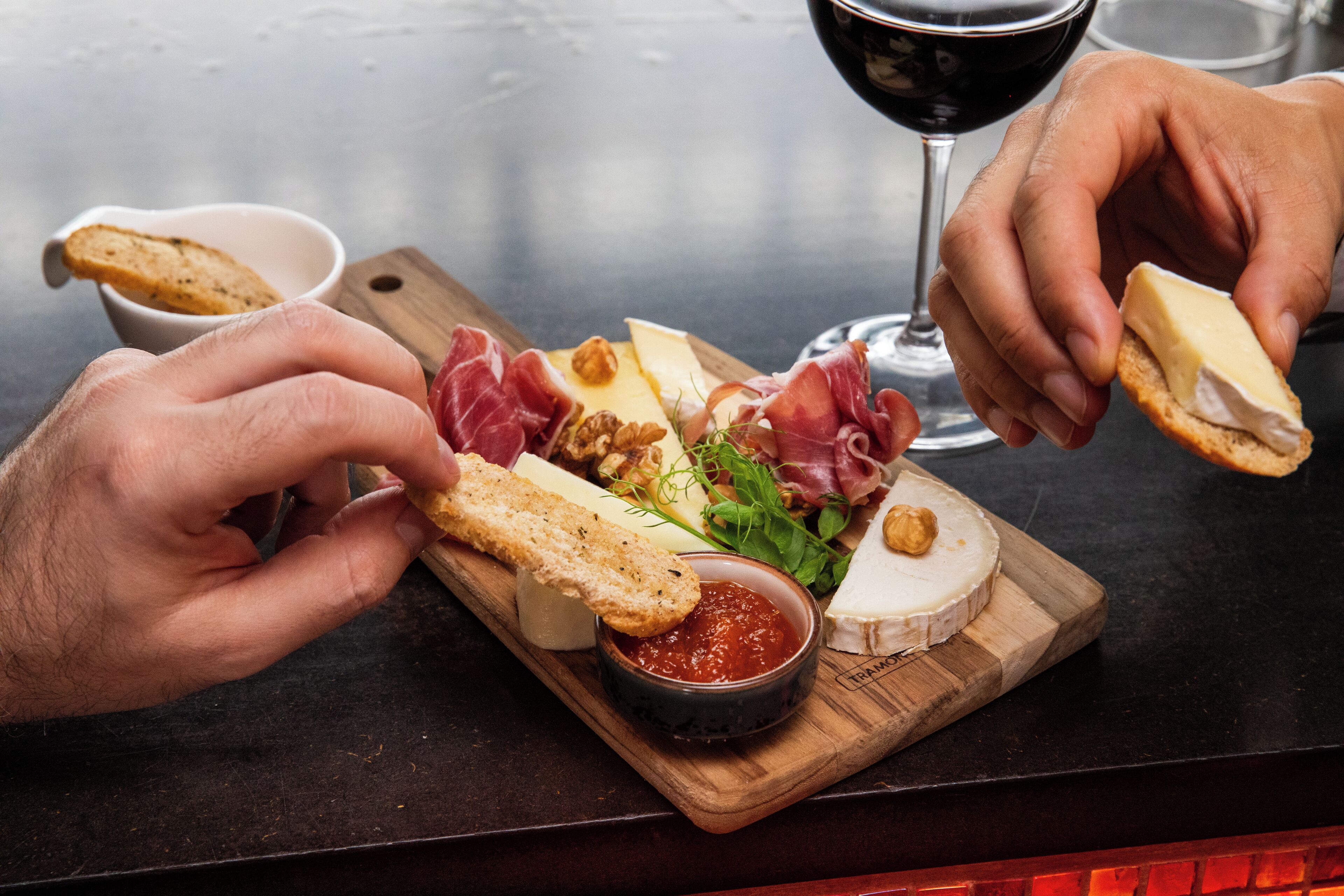 Une planche en bois garnie de différents fromages, de charcuteries et de noix, accompagnée d’un verre de vin rouge et d’un petit bol de sauce rouge.