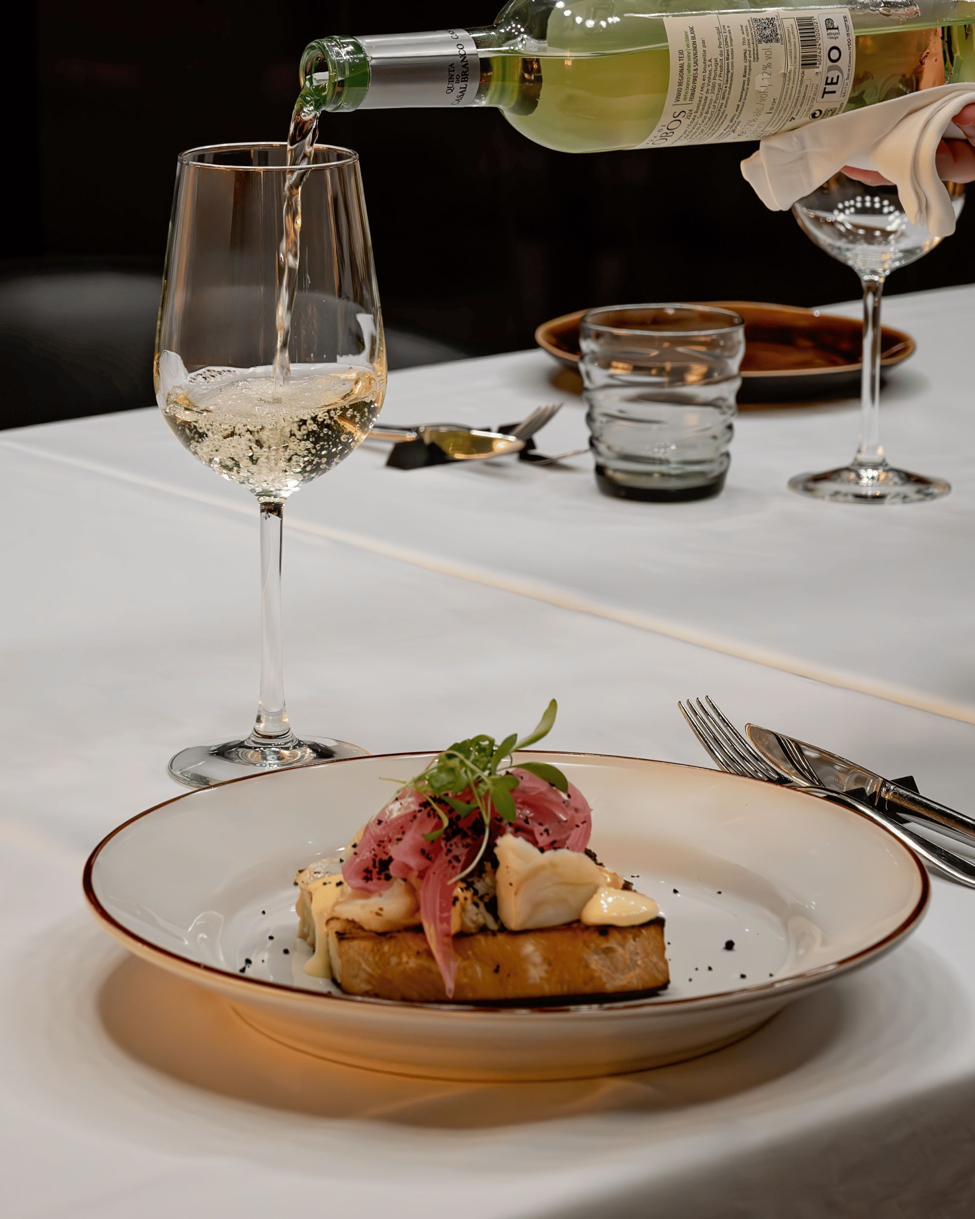 Un tavolo da pranzo con un piatto di cibo, un bicchiere di vino bianco e una bottiglia di vino che viene versata nel bicchiere.