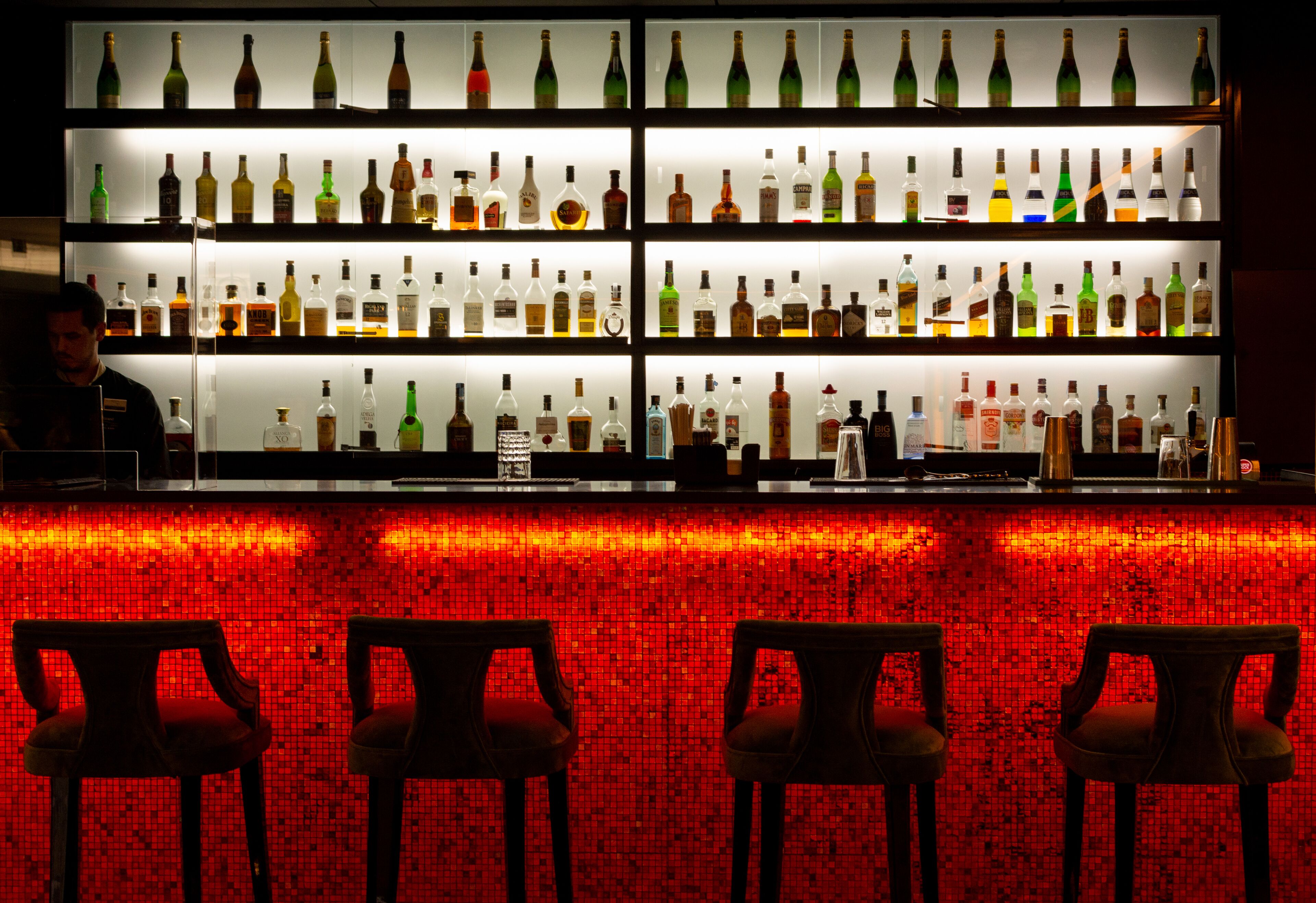 Un'area bar con scaffali multipli pieni di bottiglie, uno schienale a mosaico rosso e tre sgabelli da bar.