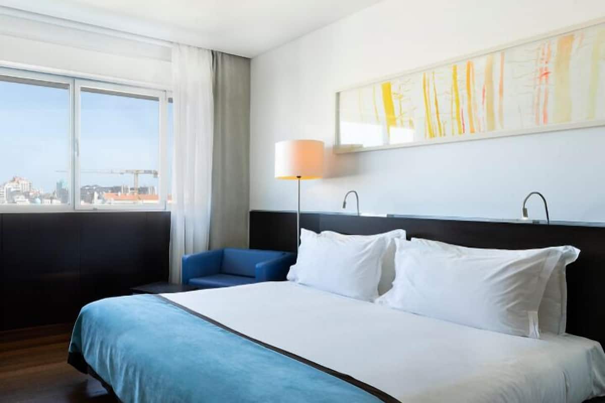 A hotel room with a large bed, a blue armchair, a floor lamp, and a city view through the window.