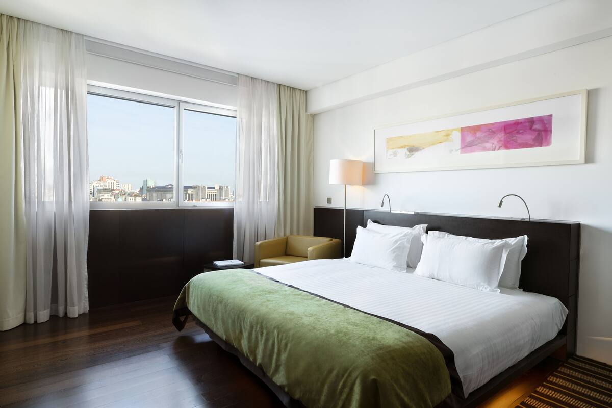 A hotel room with a large bed, a chair, a window with curtains, a lamp, and a framed picture on the wall.
