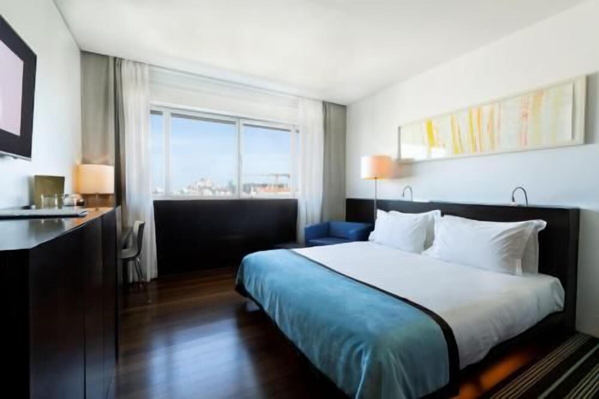 A hotel room with a bed, a desk, a chair, a window with a view, and a painting on the wall.