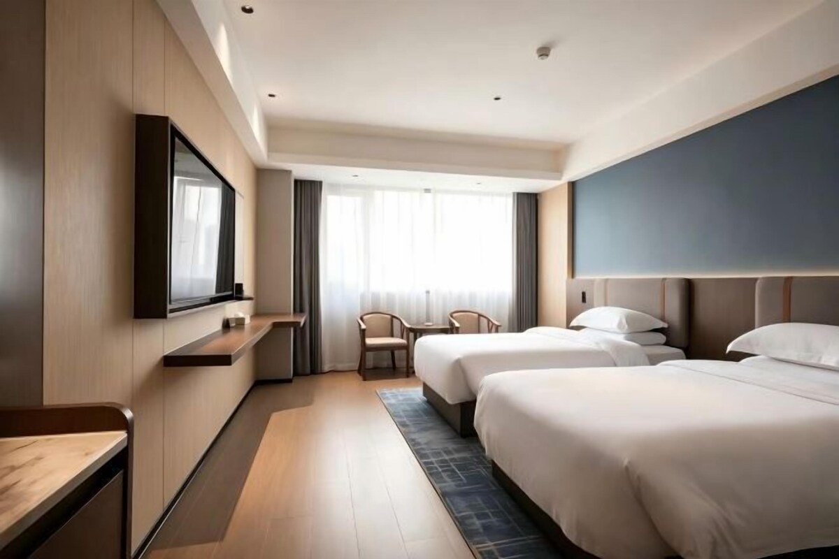 A hotel room with two beds, a TV, a desk, and a window with curtains.