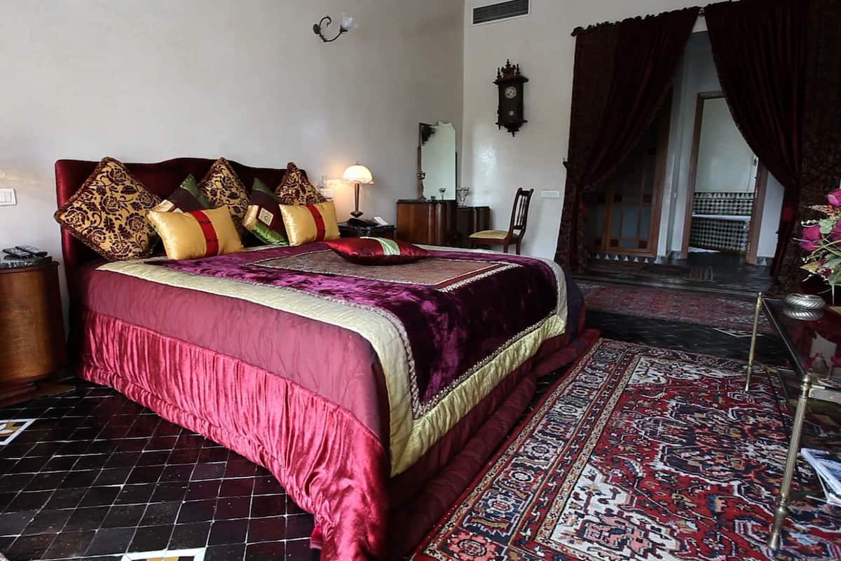 Una cama grande con cubrecama rojo y dorado, varios almohadones, dos mesitas de noche con lámparas, un espejo grande y una alfombra estampada.