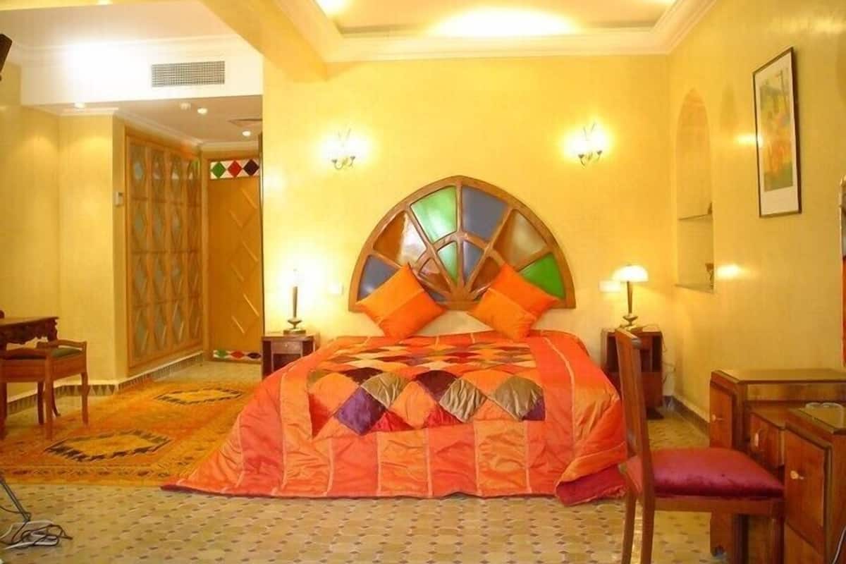 Habitación de hotel con una cama grande, un cabecero de madera, dos mesitas de noche, una butaca, un tapete estampado y una puerta con un panel decorativo.