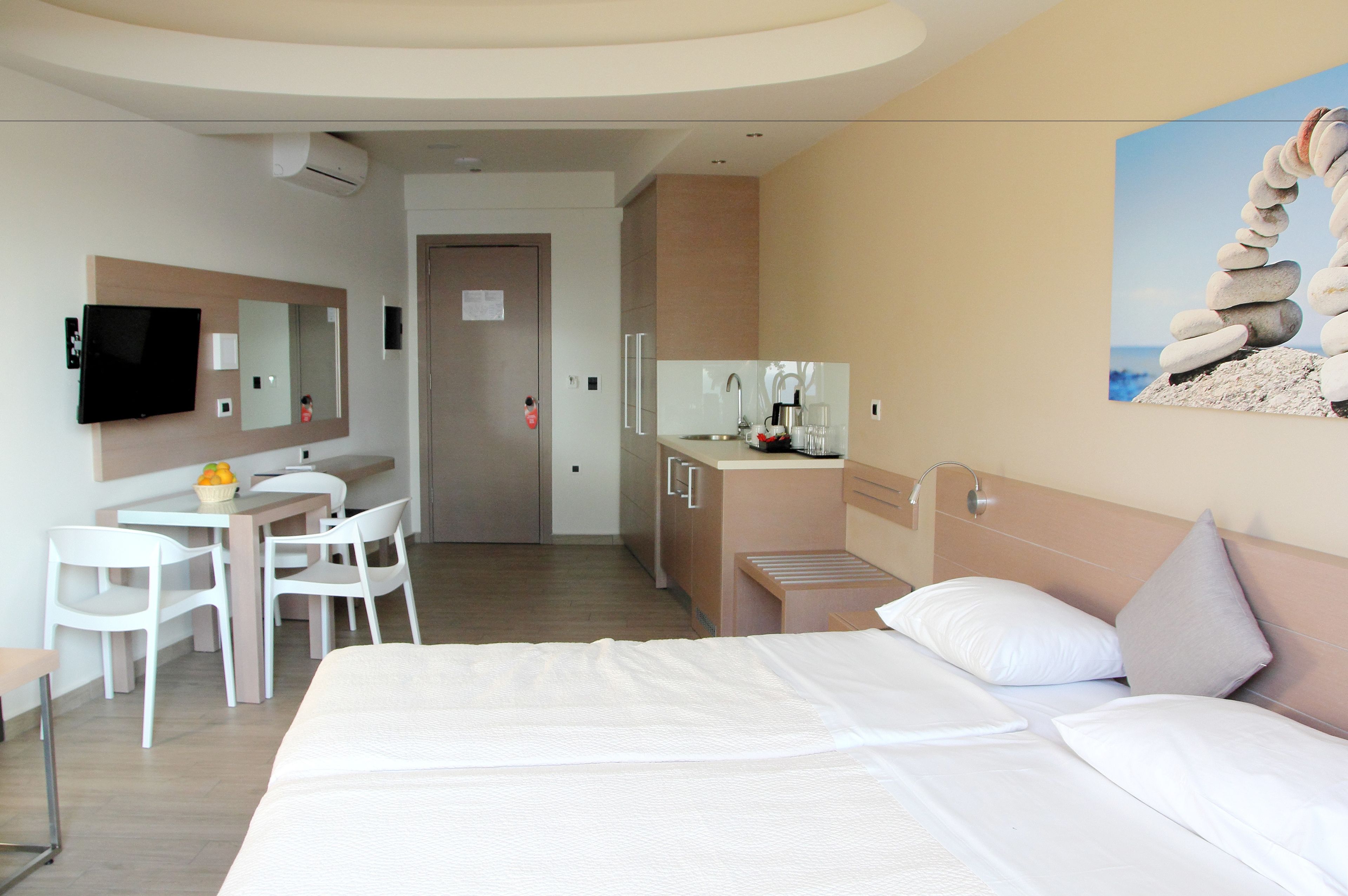 A hotel room with a bed, a desk with chairs, a TV, a mirror, a painting of stones, and a kitchenette.